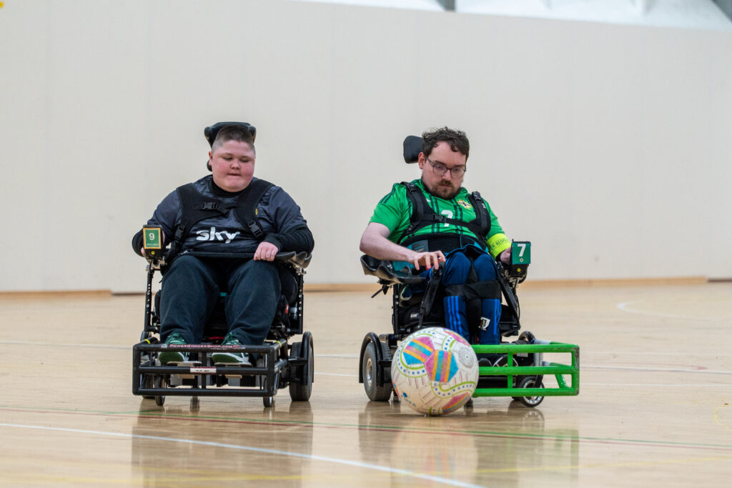 A powerchair football player driving forward with the ball during a match.